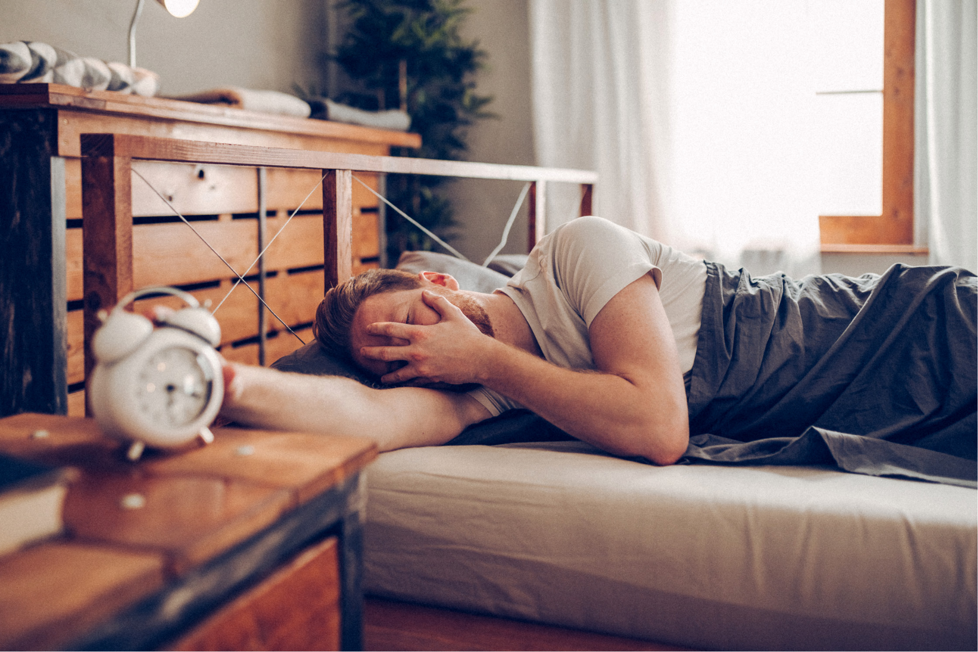 Ein Mann liegt im Bett unter einer grauen Decke und greift mit ausgestrecktem Arm nach einem weißen Wecker auf dem hölzernen Nachttisch. Mit der anderen Hand verdeckt er müde sein Gesicht vor dem hellen Licht, das durch das Fenster fällt.